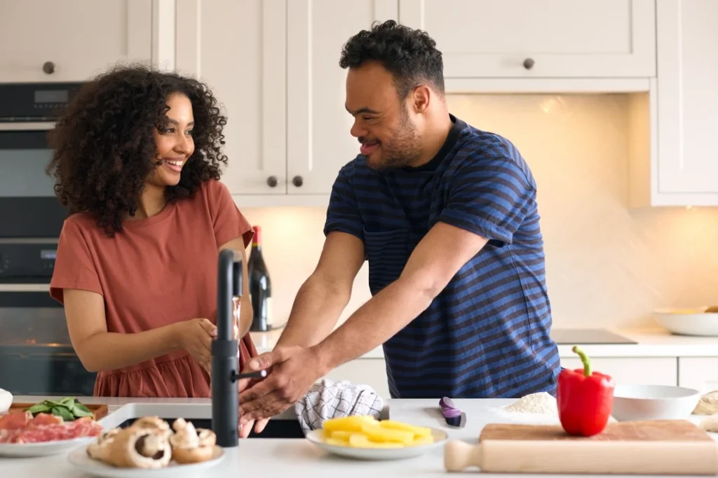 Disabled man with down syndrome washing hands in kitchen sink and smiling at support worker